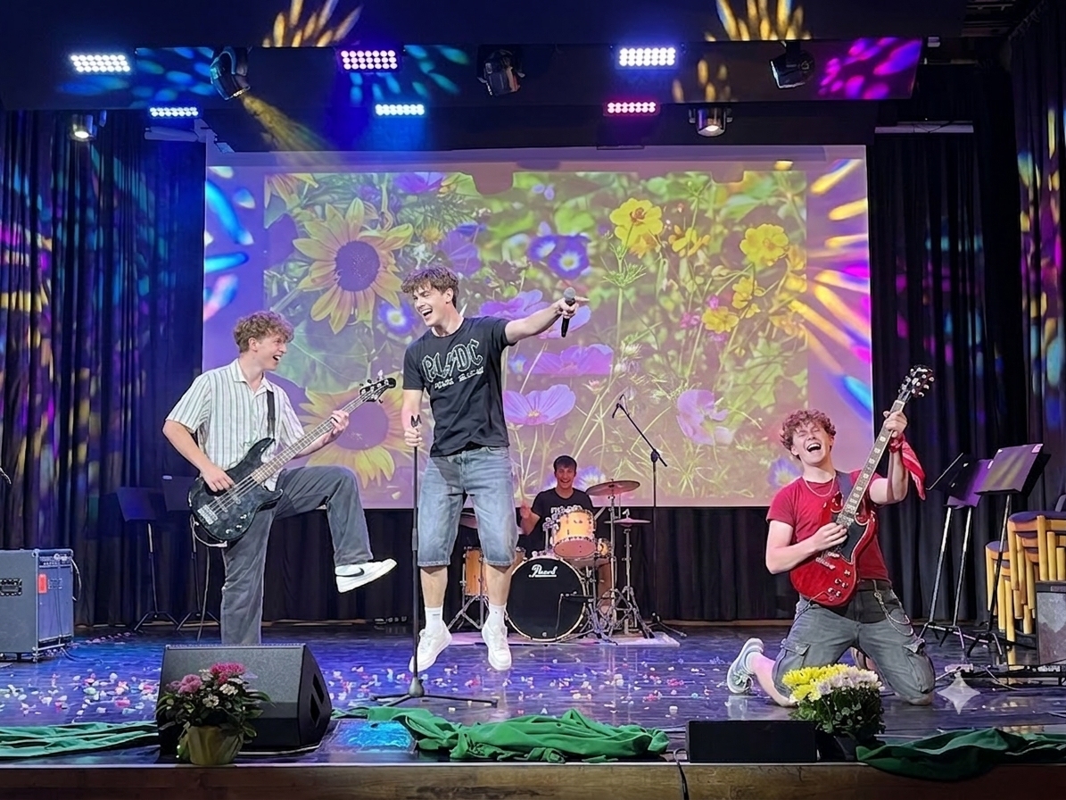 4 Musiker mit Gitarren, Schlagzeug und Mikrofone spielen Musik auf einer Bühne, Hintergrundbild ist eine Blumenwiese